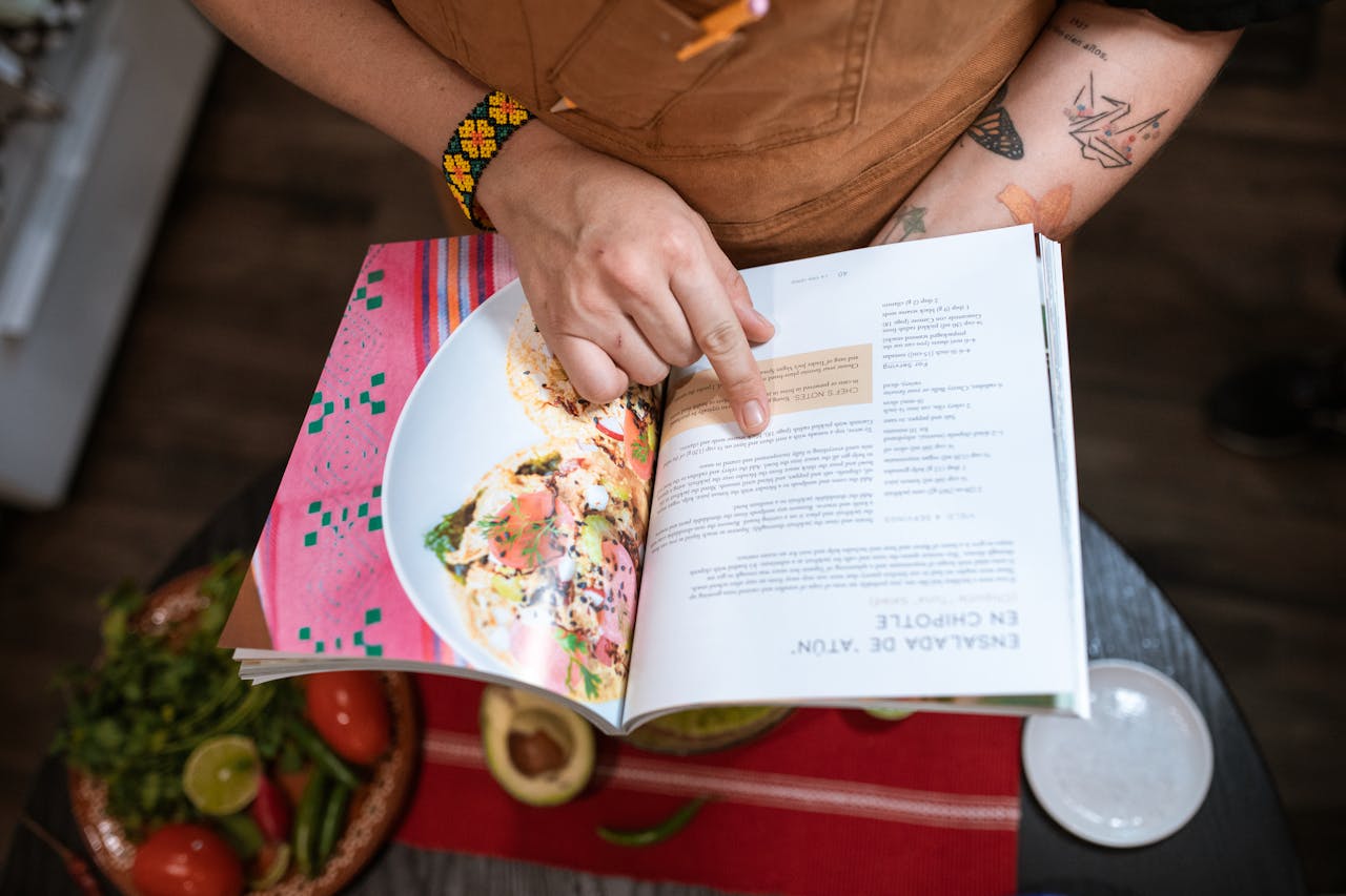services-03 Close-up of a person pointing at a recipe in an open cookbook next to fresh ingredients.