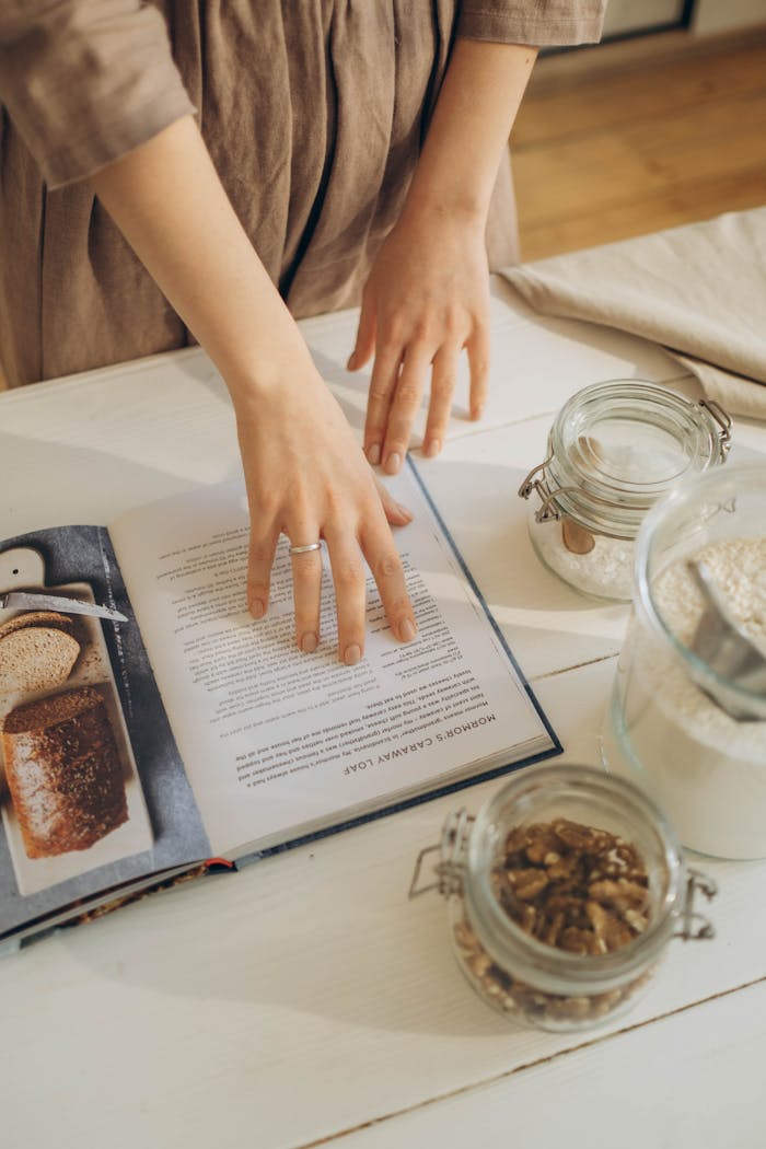 about-us A person examines a cookbook surrounded by glass jars of ingredients on a kitchen table.